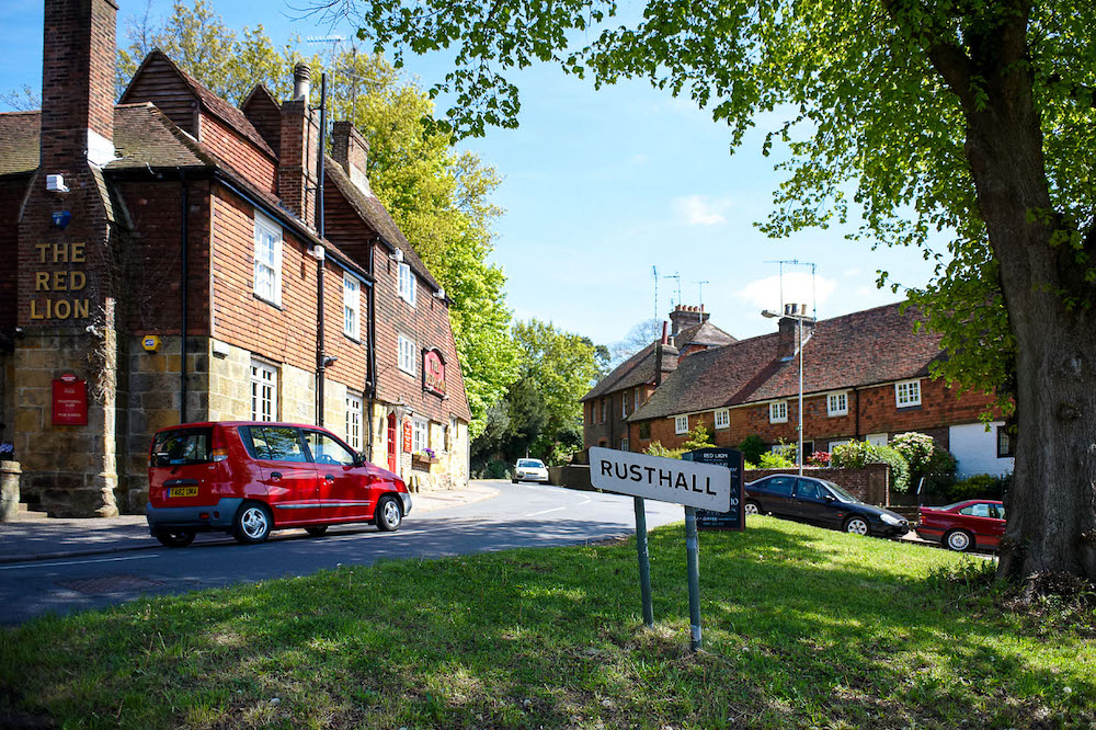 vtw-rusthall-slide-1 A street in Rusthall with greenery framing the image with the village sign of Rusthall in the centre, a pub to the left and houses to the right